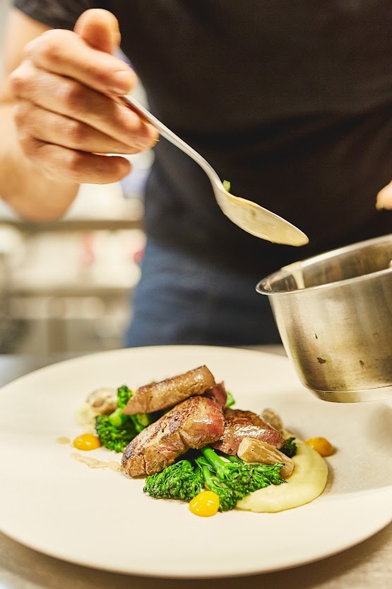 Küchenchef löffelt Sauce über Fleischgericht mit Brokkolini – handwerkliche Präzision im Restaurant Weinforum Franken